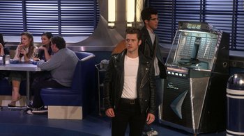 Movie still from “Grease Live!” (2016), directed by Thomas Kail – A man in a leather jacket standing in front of an arcade machine; Wide shot, Over the shoulder angle