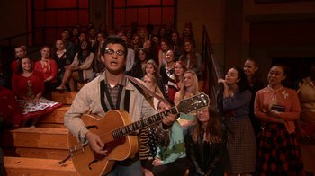Movie still from “Grease Live!” (2016), directed by Thomas Kail – A man playing a guitar in front of a group of people; Wide shot, Over the shoulder angle