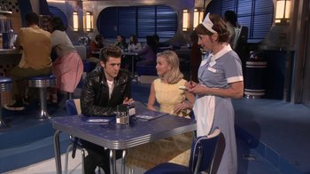 Movie still from “Grease Live!” (2016), directed by Thomas Kail – A group of people standing around a table; Medium shot, Over the shoulder angle