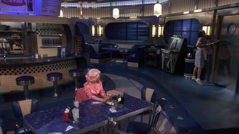 Movie still from “Grease Live!” (2016), directed by Thomas Kail – A woman sitting at a table in a restaurant; Wide shot, High angle