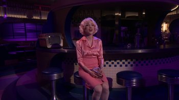 Movie still from “Grease Live!” (2016), directed by Thomas Kail – A woman in a pink dress sitting on a bar; Wide shot, Over the shoulder angle
