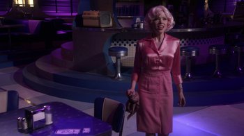 Movie still from “Grease Live!” (2016), directed by Thomas Kail – A woman in a pink dress holding a purse in a room; Medium shot, Over the shoulder angle