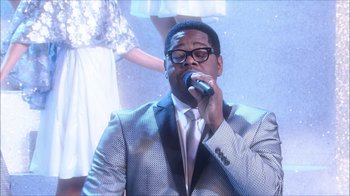 Movie still from “Grease Live!” (2016), directed by Thomas Kail – A man in a suit is singing into a microphone; Medium shot, Low angle