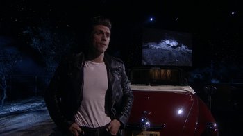 Movie still from “Grease Live!” (2016), directed by Thomas Kail – A man in a leather jacket standing in front of a car; Medium shot, Over the shoulder angle