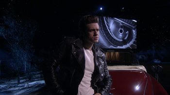 Movie still from “Grease Live!” (2016), directed by Thomas Kail – A man in a leather jacket standing in front of a car; Wide shot, Over the shoulder angle