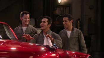 Movie still from “Grease Live!” (2016), directed by Thomas Kail – A group of men standing next to each other near a red car; Medium shot, Over the shoulder angle