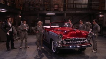 Movie still from “Grease Live!” (2016), directed by Thomas Kail – A group of people standing around a red and silver car; Wide shot, High angle