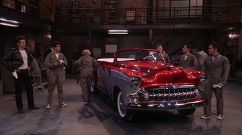 Movie still from “Grease Live!” (2016), directed by Thomas Kail – A group of people standing next to an old car; Extreme Wide shot, High angle