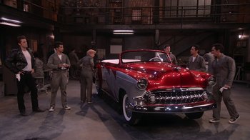 Movie still from “Grease Live!” (2016), directed by Thomas Kail – A group of people standing around a red car; Wide shot, High angle