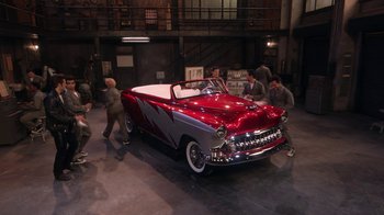 Movie still from “Grease Live!” (2016), directed by Thomas Kail – People are looking at an antique car in a museum; Extreme Wide shot, High angle