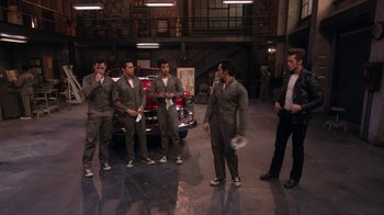 Movie still from “Grease Live!” (2016), directed by Thomas Kail – A group of men standing next to each other in front of a car; Wide shot, High angle