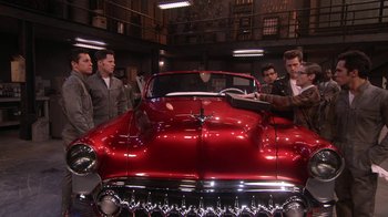 Movie still from “Grease Live!” (2016), directed by Thomas Kail – A group of men standing next to an old red car; Wide shot, High angle