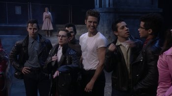 Movie still from “Grease Live!” (2016), directed by Thomas Kail – A group of young men standing next to each other on a street; Medium shot, Over the shoulder angle