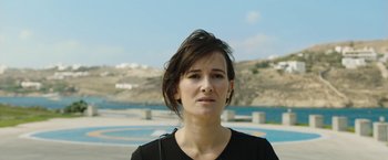 Movie still from “Greed” (2019), directed by Michael Winterbottom – A person wearing a black shirt near a body of water; Close Up shot, Low angle