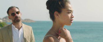 Movie still from “Greed” (2019), directed by Michael Winterbottom – A young woman wearing a white dress and a gold necklace; Close Up shot, Over the shoulder angle