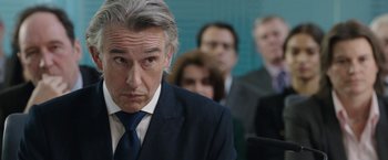 Movie still from “Greed” (2019), directed by Michael Winterbottom – A person wearing a suit and tie; Close Up shot, Over the shoulder angle