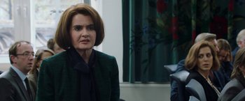 Movie still from “Greed” (2019), directed by Michael Winterbottom – A woman in a green jacket is standing in front of a window; Close Up shot, Over the shoulder angle