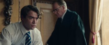 Movie still from “Greed” (2019), directed by Michael Winterbottom – A man in a suit and tie next to another man in a suit and tie; Medium shot, Over the shoulder angle