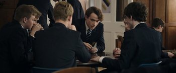 Movie still from “Greed” (2019), directed by Michael Winterbottom – A group of young men sitting around a table; Medium shot, Over the shoulder angle