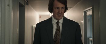 Movie still from “Greed” (2019), directed by Michael Winterbottom – A man in a suit and tie standing in a room; Medium shot, Low angle