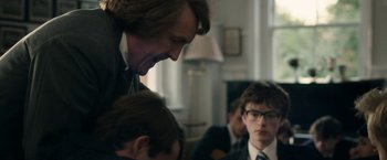 Movie still from “Greed” (2019), directed by Michael Winterbottom – A group of people sitting in a living room talking; Medium shot, Over the shoulder angle