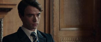 Movie still from “Greed” (2019), directed by Michael Winterbottom – A man in a suit and tie in front of a wooden wall; Close Up shot, Over the shoulder angle