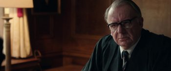 Movie still from “Greed” (2019), directed by Michael Winterbottom – An older man in a black robe and glasses; Close Up shot, Over the shoulder angle