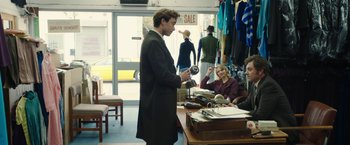 Movie still from “Greed” (2019), directed by Michael Winterbottom – A man in a black suit is looking at a camera in a store; Medium shot, Over the shoulder angle