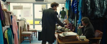 Movie still from “Greed” (2019), directed by Michael Winterbottom – A man in a black coat is shaking hands with a woman in a purple dress; Medium shot, Over the shoulder angle