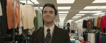 Movie still from “Greed” (2019), directed by Michael Winterbottom – A man wearing a suit and tie in a store; Medium shot, Over the shoulder angle