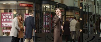 Movie still from “Greed” (2019), directed by Michael Winterbottom – A man in a brown coat and tie standing in front of a store; Medium shot, Over the shoulder angle