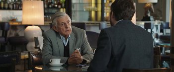 Movie still from “Greed” (2019), directed by Michael Winterbottom – A man sitting at a table talking to another man in a suit; Medium shot, Over the shoulder angle