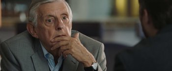 Movie still from “Greed” (2019), directed by Michael Winterbottom – An older man in a gray suit is looking at the camera; Close Up shot, Over the shoulder angle