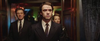 Movie still from “Greed” (2019), directed by Michael Winterbottom – A man wearing a suit and tie standing next to a woman; Medium shot, Over the shoulder angle