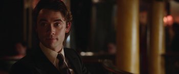 Movie still from “Greed” (2019), directed by Michael Winterbottom – A man in a suit and tie sitting in a chair; Close Up shot, Over the shoulder angle