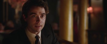 Movie still from “Greed” (2019), directed by Michael Winterbottom – A man in a suit and tie sitting at a table; Close Up shot, Over the shoulder angle