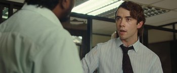 Movie still from “Greed” (2019), directed by Michael Winterbottom – A man in a tie talking to another man in an office; Close Up shot, Over the shoulder angle