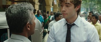Movie still from “Greed” (2019), directed by Michael Winterbottom – A man wearing a tie talking to another man in front of a crowd of onlookers; Close Up shot, Over the shoulder angle