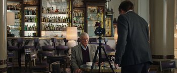Movie still from “Greed” (2019), directed by Michael Winterbottom – An older man sitting at a table in front of a camera; Medium shot, Over the shoulder angle