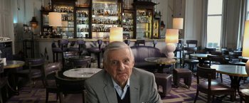 Movie still from “Greed” (2019), directed by Michael Winterbottom – An older man sitting in front of a bar; Medium shot, Low angle