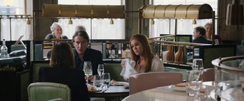 Movie still from “Greed” (2019), directed by Michael Winterbottom – A woman sitting at a table in front of two men; Medium shot, Over the shoulder angle
