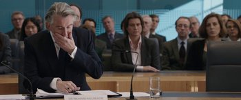 Movie still from “Greed” (2019), directed by Michael Winterbottom – A group of people sitting at a table in front of a microphone; Medium shot, Over the shoulder angle