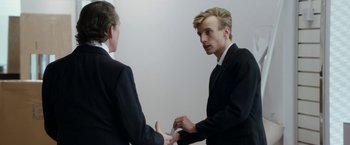 Movie still from “Greed” (2019), directed by Michael Winterbottom – A man in a suit and tie shaking hands with another man in a suit and tie; Medium shot, Over the shoulder angle