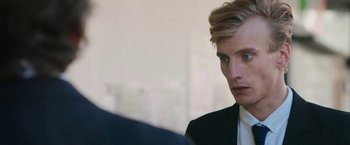 Movie still from “Greed” (2019), directed by Michael Winterbottom – A person wearing a suit and tie; Close Up shot, Over the shoulder angle