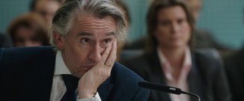 Movie still from “Greed” (2019), directed by Michael Winterbottom – A person sitting at a table holding his hand up to his face; Close Up shot, Over the shoulder angle