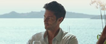 Movie still from “Greed” (2019), directed by Michael Winterbottom – A person sitting at a table holding a wine glass; Close Up shot, Over the shoulder angle