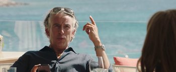 Movie still from “Greed” (2019), directed by Michael Winterbottom – A man sitting in front of the ocean pointing to the sky; Close Up shot, Low angle