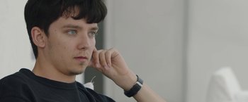 Movie still from “Greed” (2019), directed by Michael Winterbottom – A young man with blue eyes is holding his hand up to his face; Close Up shot, Over the shoulder angle