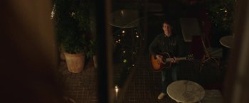 Movie still from “Greed” (2019), directed by Michael Winterbottom – A man playing a guitar in a dark room; Wide shot, Over the shoulder angle