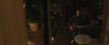 Movie still from “Greed” (2019), directed by Michael Winterbottom – A man sitting on the ground playing a guitar; Wide shot, Over the shoulder angle